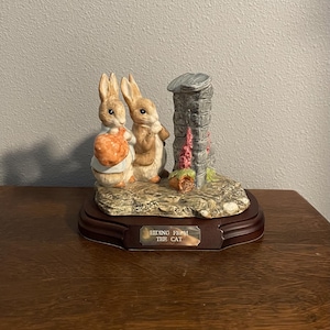 Puede incluir: Figura de cerámica con dos conejos escondidos cerca de una estructura de piedra. Un conejo sostiene una calabaza. La base de madera oscura tiene una placa que dice "Hiding From The Cat".