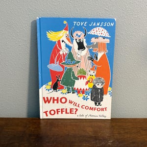 May include: A hardcover book titled "Who Will Comfort Toffle?" by Tove Jansson. The cover features colorful illustrations of whimsical characters against a blue background. The book is a tale of Moomin Valley.