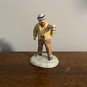 May include: A ceramic figurine of a man in a tan jacket, brown trousers, and a white hat, holding a yellow lantern. The figure stands on a white, oval base. The man has a moustache and a red scarf.