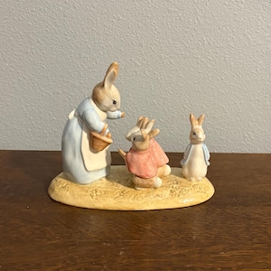 Puede incluir: Figura de cerámica que representa tres conejos en una escena pastoral. Un conejo viste un vestido y delantal azules, sosteniendo una cesta. Otro conejo lleva un abrigo rosa, y el tercero una chaqueta azul. La base es de color amarillo claro.