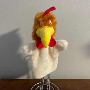 May include: A plush chicken hand puppet with a yellow beak, red comb and wattle, and orange and white feathers. The puppet is displayed on a chrome stand, showcasing its design and features.