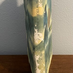 May include: The Witch of Blackbird Pond by Elizabeth George Speare. A vintage paperback book with a blue and green cover featuring a woman in a long dress and a man in a hat. The title is printed in white on the spine.