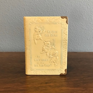 Puede incluir: Una alcancía vintage hecha de cuero sintético crema con ribete dorado. La parte delantera presenta tres cerdos en relieve con el texto "We Save Our Coins - So Who's Afraid of the Big Bad Wolf!"