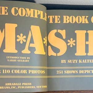 Op de afbeelding: Open boek met de titel "THE COMPLETE BOOK OF M*A*S*H" met gele letters op een teal achtergrond. Het boek bevat de naam van de auteur, een inleiding door Larry Gelbart en vermeldt meer dan 110 kleurenfoto's en 251 afgebeelde shows.