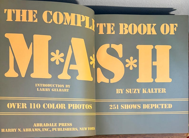Op de afbeelding: Open boek met de titel "THE COMPLETE BOOK OF M*A*S*H" met gele letters op een teal achtergrond. Het boek bevat de naam van de auteur, een inleiding door Larry Gelbart en vermeldt meer dan 110 kleurenfoto's en 251 afgebeelde shows.