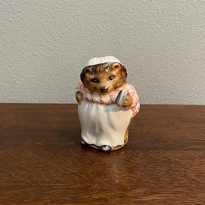 May include: A ceramic figurine of a brown and white hedgehog wearing a red and white checkered apron and holding a small silver object. The figurine is standing on a wooden surface.