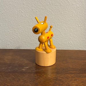 May include: A wooden toy dog with a yellow body, black eyes, and red spots. The dog has a cylindrical head and is mounted on a small, round wooden base. The toy is a vintage style and is made of wood.