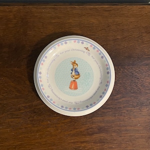 Puede incluir: Un plato de cerámica blanco con un borde azul y rosa. El centro del plato presenta una ilustración azul y blanca de Peter Rabbit de pie sobre un taburete rojo y blanco. El plato está decorado con el texto "For your christening" repetido alrededor del borde.