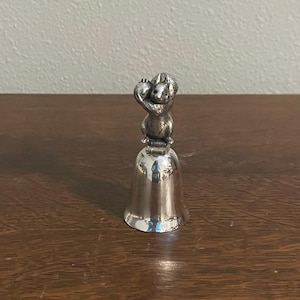 May include: A silver bell with a squirrel figurine on top holding an acorn. The squirrel is sitting on the bell's handle.