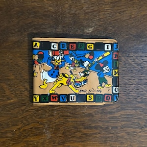 Peut inclure: Portefeuille pliable beige avec des personnages Disney et l'alphabet. Donald Duck, Mickey Mouse, Pluto et Dingo sont représentés dans des illustrations colorées. Le portefeuille est de forme rectangulaire et est fait d'un matériau de couleur claire.