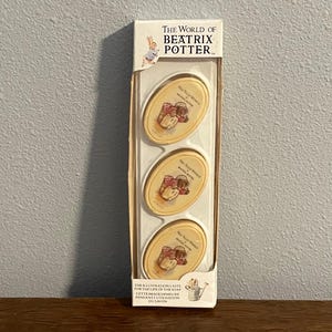 Puede incluir: Un juego de tres jabones ovalados con ilustraciones de "The World of Beatrix Potter". Cada jabón tiene un fondo crema con una imagen detallada de una cesta de frutas. El embalaje incluye texto.