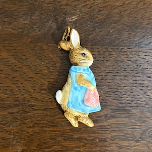 Puede incluir: Una figura de conejo de cerámica, que presenta un conejo marrón claro con un delantal azul y una bolsa rosa. El conejo tiene una cola blanca y está sobre un fondo de madera. La figura es probablemente un artículo decorativo.