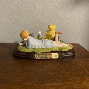 May include: A ceramic figurine depicting a summer picnic scene. The figurine features Winnie the Pooh, Piglet, and Christopher Robin. The base is a dark brown color, and the text "SUMMERS DAY PICNIC" is displayed on a gold plaque.