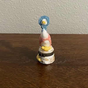 Puede incluir: Figura de cerámica de un ganso con un gorro azul y un vestido rosa y blanco. El ganso está posado sobre una pequeña caja redonda con tapa con bisagras. La caja está decorada con detalles dorados y pollitos amarillos.