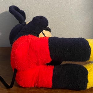May include: A plush toy of a famous cartoon mouse, lying on its side. The toy features a black body, red shorts, yellow feet, and large black ears. The tail is black and curved. The toy is made of a soft, fuzzy material.