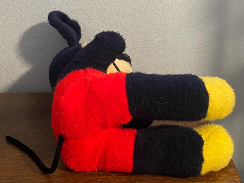 May include: A plush toy of a famous cartoon mouse, lying on its side. The toy features a black body, red shorts, yellow feet, and large black ears. The tail is black and curved. The toy is made of a soft, fuzzy material.