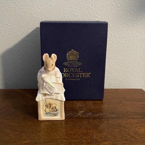 Puede incluir: Figura de Royal Worcester de un ratón con vestido y delantal, sosteniendo un plumero y un bloc de notas. Frente a una caja azul marino con letras doradas y un escudo. La figura está sobre una superficie de madera.