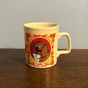 Könnte beinhalten: Eine cremefarbene Keramik-Tasse mit einer Comicfigur, die Noten hält. Die Figur ist braun mit rundem Gesicht, in einem roten und orangefarbenen ovalen Rahmen. Die Tasse hat einen gebogenen Henkel und orangefarbene Blumenmuster.