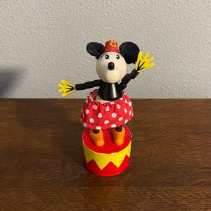 May include: A vintage wooden puppet of Mickey Mouse dressed in a red and white polka dot skirt and a red hat with a yellow flower. The puppet is standing on a red and yellow stage.