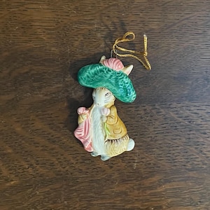 Può includere: Statuina in ceramica di un coniglio con cappello verde e cappotto giallo, che tiene un panno rosa. Il coniglio ha una faccia bianca e una coda soffice. Un filo d'oro è attaccato per appenderlo. Articolo decorativo.