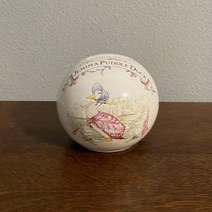 May include: A round, white ceramic Jemima Puddle-Duck bank. The bank features a detailed illustration of Jemima Puddle-Duck in a bonnet and shawl, standing in a grassy field. The words "Jemima Puddle-Duck" are written in a banner around the top.