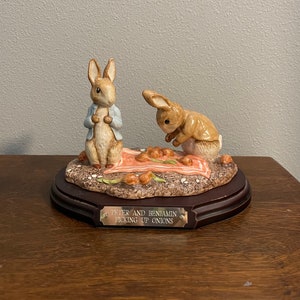 Puede incluir: Conjunto de figuras de Peter Rabbit y Benjamin Bunny. Los dos conejos están hechos de cerámica y están pintados en colores realistas. Peter está de pie, mientras que Benjamin está agachado, recogiendo cebollas. Las figuras están sobre una base de madera con una placa que dice "Peter y Benjamin recogiendo cebollas".