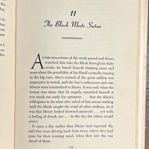 May include: Open book page with text. The title "The Black Meets Satan" is displayed in elegant script above a horizontal line. The text is in black ink on aged, off-white paper, with a large initial letter.