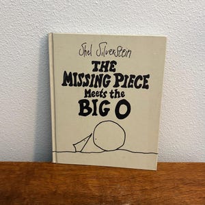 May include: A hardcover book titled "The Missing Piece Meets the Big O" by Shel Silverstein. The book has a light beige cover with black lettering and a simple illustration of a circle and a triangle. The book is resting on a wooden surface.