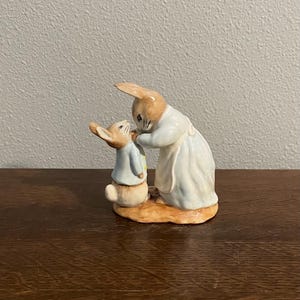 May include: A ceramic figurine depicting two rabbits. One rabbit, wearing a blue jacket, is being attended to by another rabbit in a white dress and apron. The figurine is painted in soft, muted colors and sits on a brown base.