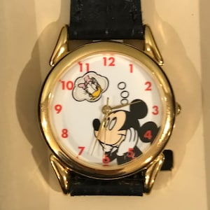 Puede incluir: Un reloj Mickey Mouse dorado con una correa de cuero negro. La esfera del reloj presenta a Mickey Mouse mirando hacia arriba a una burbuja de pensamiento con Donald Duck dentro. Los números del 1 al 12 están en rojo.