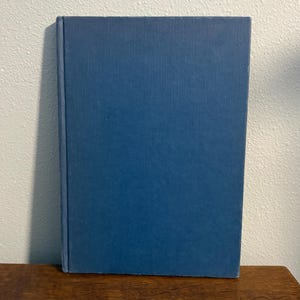 Puede incluir: Un libro de tapa dura con una cubierta azul texturizada. El libro está de pie sobre una superficie de madera. La cubierta es de color azul medio sólido y el libro parece estar en buenas condiciones.