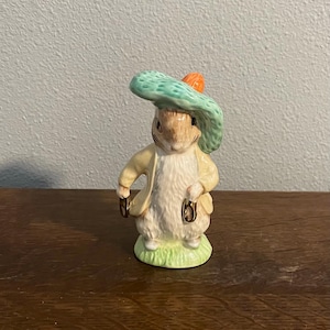 Puede incluir: Figura de cerámica de un conejo con sombrero verde claro con un detalle naranja y abrigo amarillo. El conejo está de pie sobre un trozo de hierba verde, sosteniendo una pequeña bolsa en cada mano. Un objeto de colección.