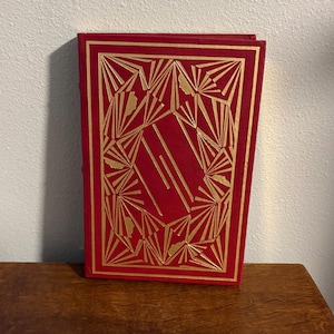 Puede incluir: Un libro rojo con un diseño geométrico dorado en la portada. El diseño presenta una forma rectangular central rodeada de líneas y formas radiales. El libro está sobre una superficie de madera.