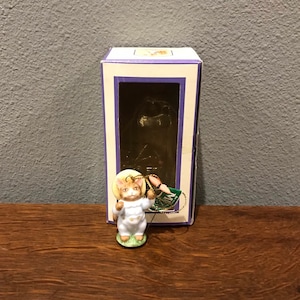 Puede incluir: Una figurita de porcelana de un gato con un atuendo blanco y azul y un sombrero de paja. El gato sostiene un globo rosa y está de pie sobre una base verde. La figurita está empaquetada en una caja púrpura y blanca con una ventana transparente.