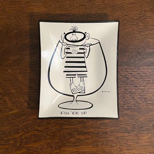 May include: A white ceramic dish with a black rim features a black and white illustration of a cartoon character holding onto the rim of a wine glass. The text "Fill 'er Up" is printed below the glass.