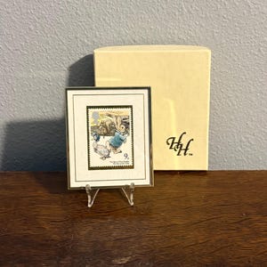 May include: A framed postage stamp featuring a rabbit and duck illustration, displayed on a clear stand. The stamp is set against a white background with a gold frame. A beige box with the initials "HH" is behind the frame.