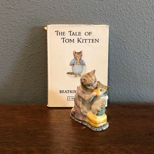 Puede incluir: Una figurita de porcelana de una gata madre alimentando a un gatito de un cuenco. La figurita está en un esquema de colores azul claro, rosa y amarillo. La figurita está al lado de un libro titulado "The Tale of Tom Kitten" de Beatrix Potter.
