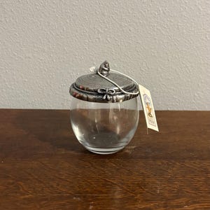May include: A clear glass jar with a silver-colored lid and a small tag. The lid has a decorative design and a small handle. The jar is sitting on a wooden surface.