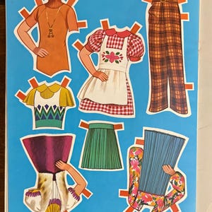 May include: A collection of paper doll clothing items on a blue background, including an orange turtleneck, a red and white gingham dress with an apron, brown plaid trousers, a yellow and green top, a green pleated skirt, and floral outfits.