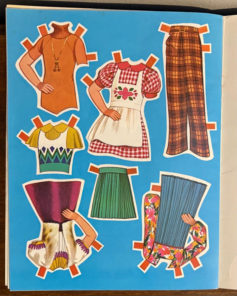 May include: A collection of paper doll clothing items on a blue background, including an orange turtleneck, a red and white gingham dress with an apron, brown plaid trousers, a yellow and green top, a green pleated skirt, and floral outfits.