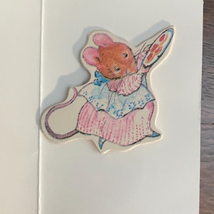 May include: A vintage greeting card featuring a charming illustration of a mouse holding a pie. The mouse is dressed in a pink and blue dress with a matching bow. The card is open, revealing a blank interior.