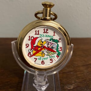 May include: Gold pocket watch with a Winnie the Pooh design. The watch face features Winnie the Pooh wearing a Santa hat and holding a gift. The text on the watch face reads "It's Xmas all year long".