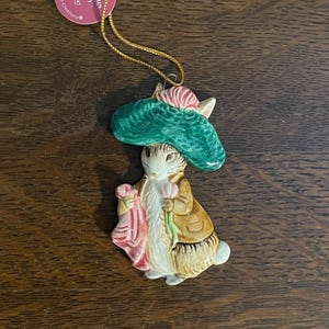 Può includere: Un ornamento in ceramica di un coniglio che indossa un cappello verde e tiene un fiore rosa. L'ornamento è etichettato "Benjamin Bunny" e fa parte di una collezione chiamata "Potter Creations".