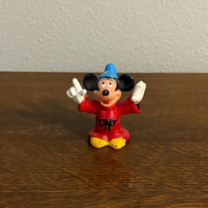 May include: A small Mickey Mouse figurine in a sorcerer's outfit. The figure is wearing a red robe, a blue hat with a gold star, and yellow shoes. Mickey has his arms raised, wearing white gloves, and a happy expression.