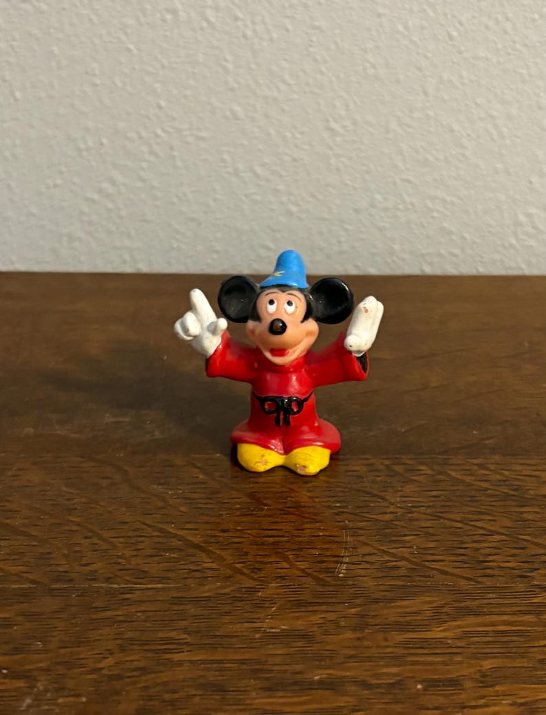 May include: A small Mickey Mouse figurine in a sorcerer's outfit. The figure is wearing a red robe, a blue hat with a gold star, and yellow shoes. Mickey has his arms raised, wearing white gloves, and a happy expression.