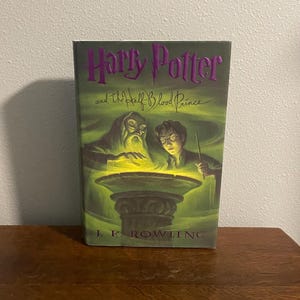 May include: A green and purple hardcover book with the title "Harry Potter and the Half-Blood Prince" by J.K. Rowling. The cover features an illustration of Harry Potter and Albus Dumbledore performing magic.