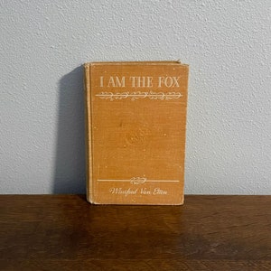 Puede incluir: Un libro de bolsillo vintage con una cubierta marrón y texto blanco que dice "I Am The Fox" de Winifred Van Etten.