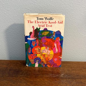May include: A vintage copy of Tom Wolfe's "The Electric Kool-Aid Acid Test". The book's cover displays a psychedelic illustration in vivid colours, including red, blue, green, and orange. The book is set against a light blue background and rests on a wooden surface.