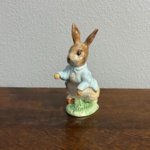 May include: A ceramic rabbit figurine, dressed in a light blue jacket and white shirt. The rabbit is brown and white with long ears, standing on a patch of green grass. A collectible item, likely from a children's story.