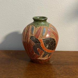 May include: A round ceramic vase featuring a textured surface with a carved turtle motif. The design incorporates shades of terracotta, black, and green, with stylized leaf patterns surrounding the central turtle figure and a green rim.